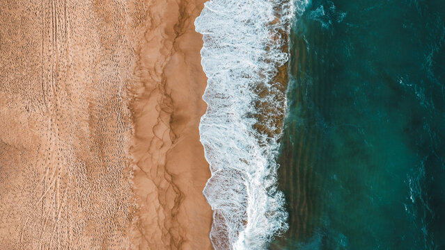 Aerial view of the sandy beach with turquoise ocean waves crashing against the shore, showing footprints in the sand and clear water Nazare, Leiria District, Portugal.