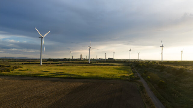 Aerial view of a wind farm with multiple turbines set in green and brown agricultural fields under a dramatic cloudy sky during golden hour in Frodsham, England, United Kingdom.
