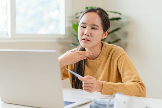 Sick Asian woman consulting doctor online via laptop video call holding thermometer and touching her sore throat at home telemedicine health care telehealth remote medical counseling concept