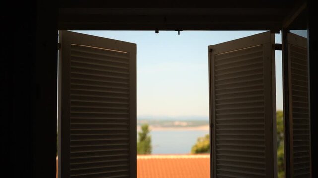 View from inside a room looking through open white louvered shutters toward a coastal sea view with terracotta roof tiles and Mediterranean greenery. 4K.