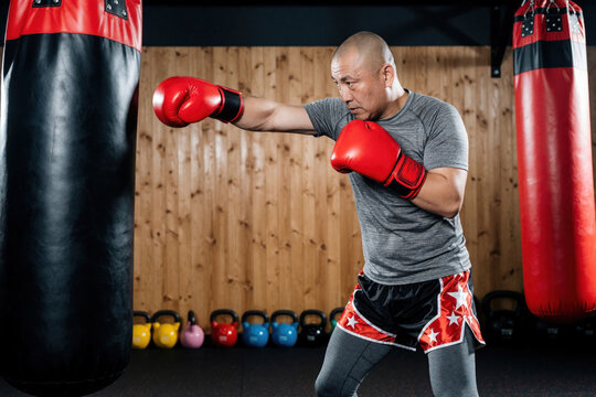 Man throws a punch at punching bag in gym. Bald man wearing red gloves boxing in athletic center. Body builder does cardio in fitness club. Man prepares for boxing sport in gym facility.