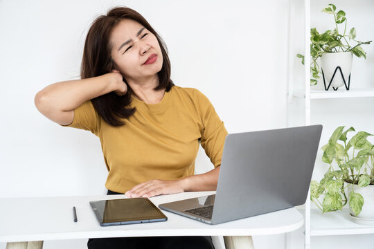 Tired Asian business woman touching neck suffering from neck pain, office syndrome, and muscle strain while working on laptop computer at desk. Stressed female worker feeling body ache and fatigue