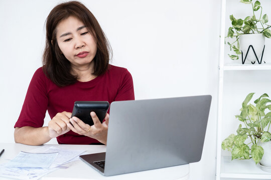 Worried Asian woman using calculator to calculate high expenses, bills, and tax at desk with laptop. Stressed female worker feeling financial problem, debt, and budget crisis in office