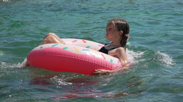 Girl floating swimming, cheerful child enjoying summer vacation playing on pink donut ring in crystal clear sea water during sunny day at beach