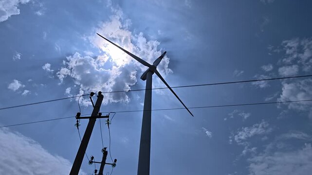 Low angle view of a wind turbine spinning and a power pole silhouetted against a sunny blue sky