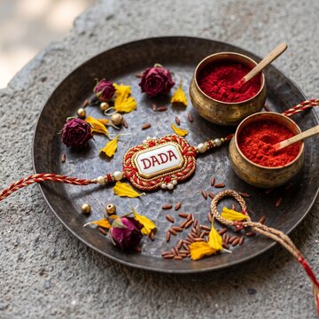 A traditional Rakhi ceremony setup featuring a decorative 'Dada' bracelet, brass bowls filled with red vermilion powder, scattered rose petals, and grains on a rustic metal plate.