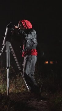 Outdoor observer in hood jacket pants focuses attention. Tripod mounted scope remains fixed as person looks. Grassy terrain extends into background where scattered points