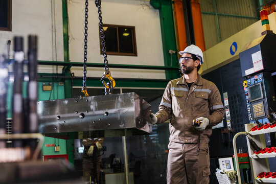 Industrial worker wearing protective equipment inspects a heavy metal component suspended by chains during a lifting operation inside a manufacturing facility.