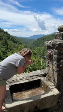 Woman drinks fresh water from a mountain fountain, then helps her dog drink. A heartwarming outdoor moment showing care, companionship and refreshment during a nature hike.