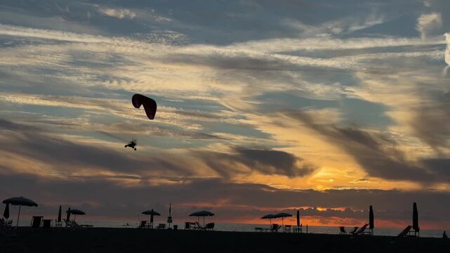 Powered paraglider pilot flying above the sea during a dramatic sunset. Golden light, colorful clouds and calm water create a scenic coastal landscape.