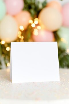 Blank white folded greeting card mock up standing upright on a festive table with pastel confetti and colorful balloons in the background, ideal for custom designs or messages