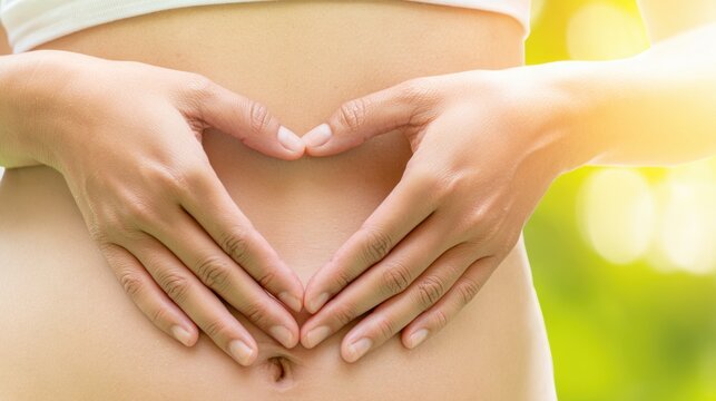Female hands forming a heart shape around the navel on a bare abdomen, symbolizing wellness and self-care in a natural outdoor setting with soft sunlight