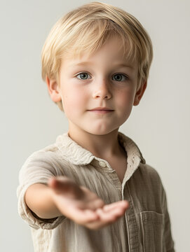 An adorable young blonde boy with innocent eyes wearing a beige linen shirt stands in a minimalist studio extending his open palm forward in a friendly gesture.