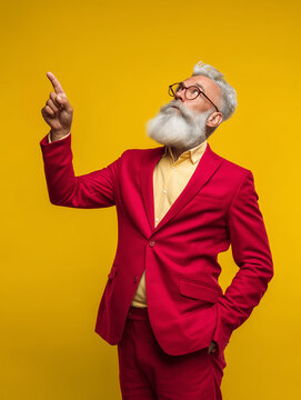 Fashionable senior man with white beard and glasses wearing a bright red suit pointing upwards against a vibrant yellow studio backdrop
