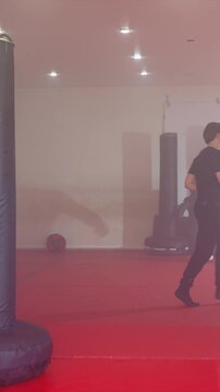 Two young men practice martial arts in indoor gym. One performs solo movements while other engages him. Environment includes black cylindrical pads torso shaped dummies
