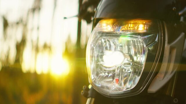 Close-up of a modern motorcycle headlight illuminated at sunset, highlighting reflective glass details and sleek design against a blurred forest background