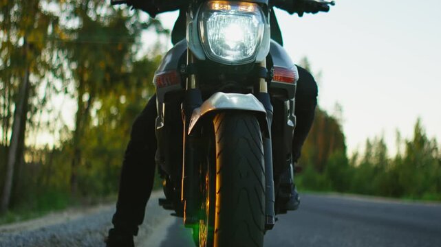 Lone motorcyclist in black helmet and gear waits on an empty forest road at sunset, front wheel and reflective visor framed by soft golden evening light