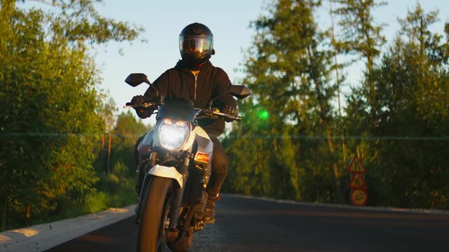 Motorcyclist riding along empty forest road at golden hour, wearing safety gear and visor helmet while cruising on modern street bike through tranquil nature