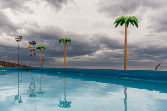 Palme artificiali attorno a una piscina fronte mare sotto cielo nuvoloso. Scena costiera pop e fuori stagione ideale per temi turismo, paesaggi marini e contrasto natura‑artificio.