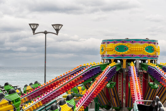 Giostra colorata con luci accese vicino al mare sotto cielo nuvoloso. Scena costiera pop ideale per temi turismo, emozioni, paesaggi marini e contrasto tra colori vivaci e atmosfera grigia.