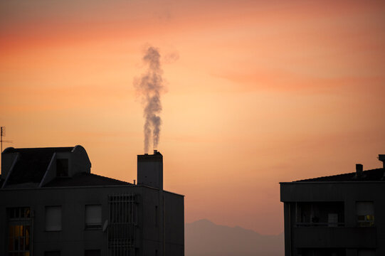 Edifici in silhouette al tramonto con camino che rilascia fumo verticale. Scena urbana calda e atmosferica, ideale per temi ambiente, citt&agrave;, inquinamento, paesaggio urbano e fotografia editoriale.