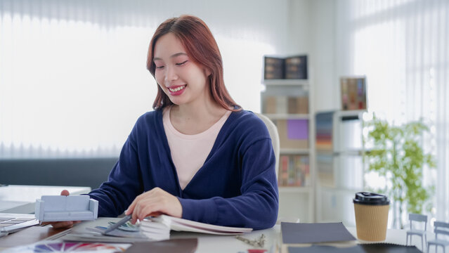 Young Asian furniture designer reviewing color swatches and material samples at modern office desk with natural light
