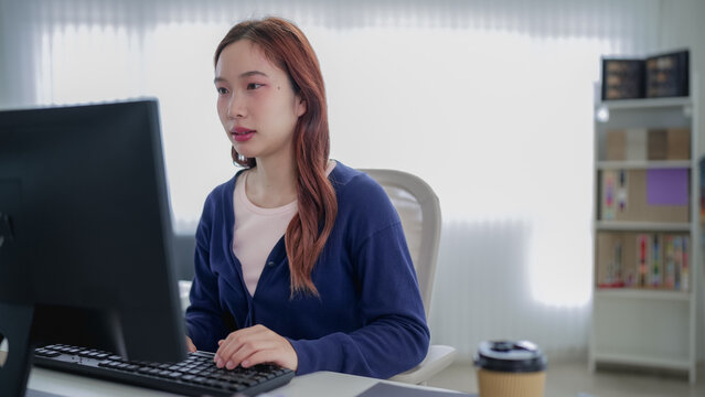 Young Asian professional working on computer in bright modern home office with bookshelf