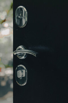 Close-up of a sleek black modern door featuring a curved chrome handle, a top keyhole, and a bottom lock, set against a bright blurred background