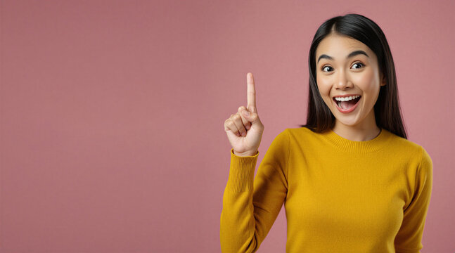 Young asian woman wearing yellow sweater points finger up. Cheerful girl smiles wide showing excitement. Female person demonstrates index gesture on pink background. Pretty model looks happy finds
