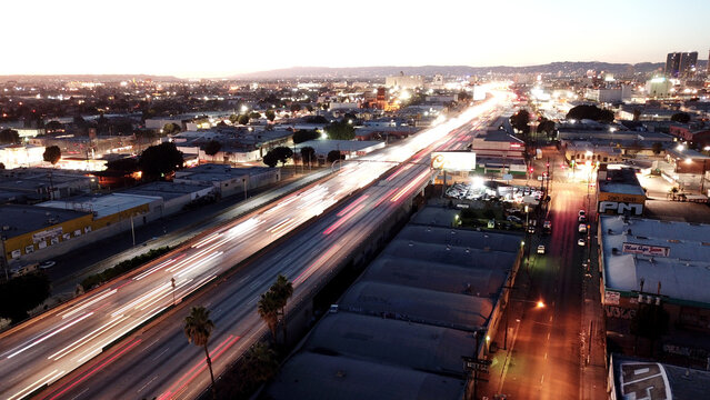 Aerial view of a busy highway with long exposure light trails from moving vehicles at dusk, surrounded by urban industrial buildings and palm trees in Los Angeles, California, United States.