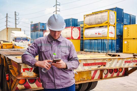 Male industrial worker using smartphone and holding water bottle by cargo truck in shipping container yard