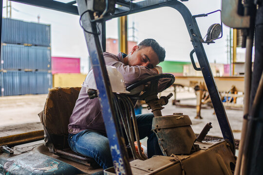 Exhausted logistics worker sleeping on forklift steering wheel in freight yard cargo container depot