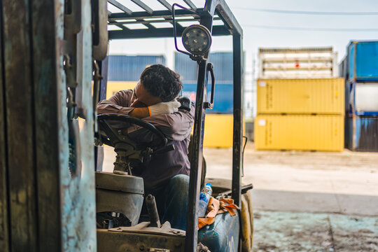 Exhausted logistics worker sleeping on forklift steering wheel in freight yard cargo container depot
