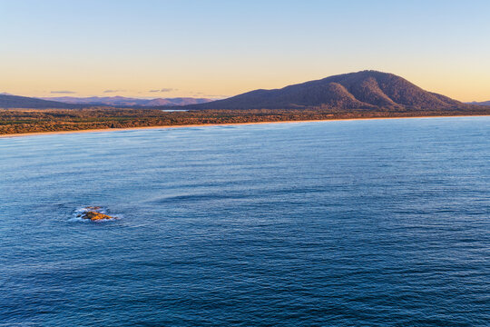 Aerial view of Diamond Beach and the surrounding Pacific Ocean, with waves rolling towards the sandy shoreline and a scenic mountain landscape rising in the background on the Mid North Coast