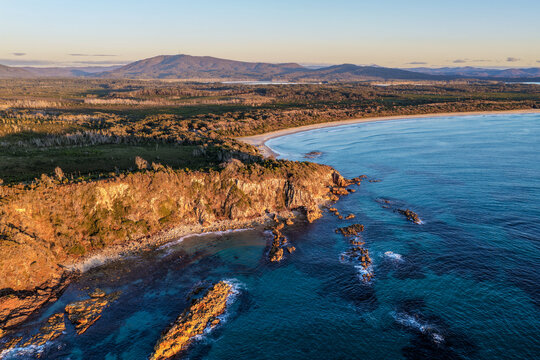 Aerial view of Diamond Beach and the surrounding Pacific Ocean, featuring a rugged rocky headland, waves rolling towards the sandy shoreline and a scenic mountain landscape 