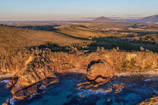 Aerial view of Diamond Head and the surrounding Pacific Ocean, featuring a rugged rocky headland, a scenic mountain landscape rising in the background on the Mid North Coast of NSW, Australia.