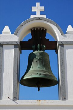 Large bronze church bell hangs in white bell tower against clear blue sky. Religious artifact has ornate carvings and a cross at top. Sacred sound maker awaits to chime.