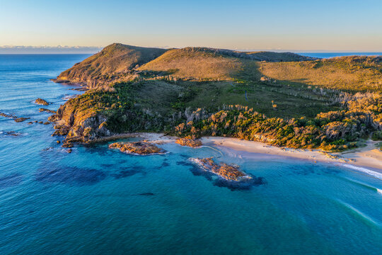 Aerial view of the rugged rocky headland at Diamond Head surrounded by clear turquoise ocean water and a sandy beach, showcasing the pristine coastline and natural coastal landscape