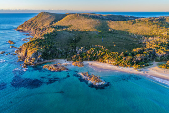 Aerial view of the rugged rocky headland at Diamond Head surrounded by clear turquoise ocean water and a sandy beach, showcasing the pristine coastline and natural coastal landscape