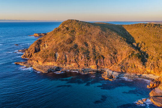 Aerial view of Indian Head surrounded by the blue waters of the Pacific Ocean, featuring a rugged rocky coastline, native coastal vegetation and waves breaking along the shoreline near Diamond Head