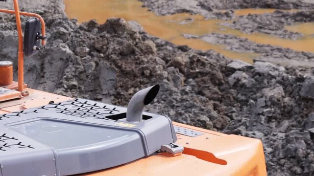 Orange-and-gray excavator housing features a curved exhaust pipe and patterned ventilation grilles; rugged industrial design contrasts with wet, clumpy mud and ochre puddles in the background
