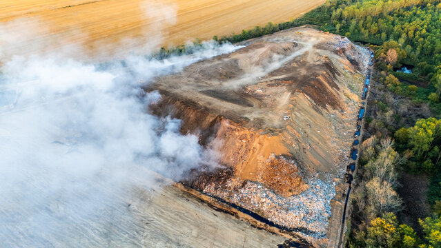 Sremska Mitrovica, Serbia - 11 August 2026: Aerial view of a large landfill site with thick smoke rising from burning waste, surrounded by agricultural fields and dense forest.