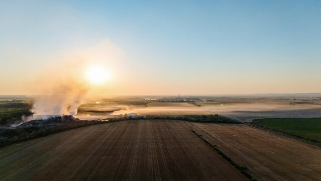 Sremska Mitrovica, Serbia - 11 August 2026: Aerial view of smoke rising from a field during sunset, with agricultural land and hazy horizon in the background.