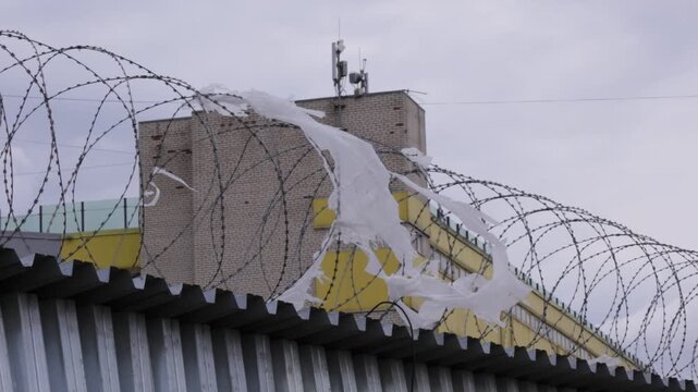 Razor wire coils top corrugated metal fence in foreground, tangled with torn white plastic strips