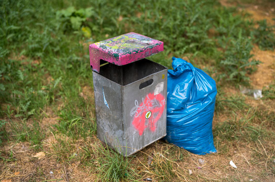 Trash bin with graffiti next to a blue garbage bag in a grassy area during the day