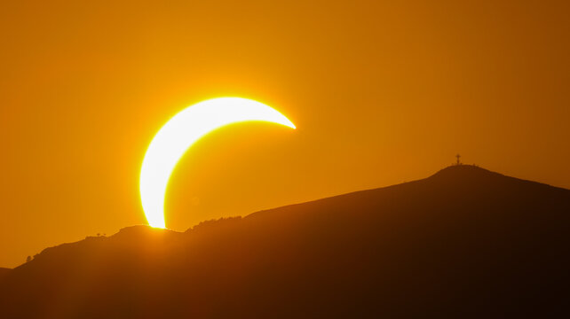 Geneva, Switzerland - 12 August 2026: View of a partial solar eclipse setting behind a mountain silhouette with a cross on the summit.