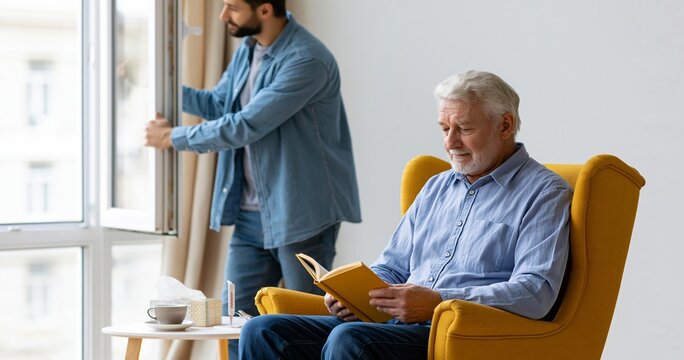 Senior man reading book while younger man opens window