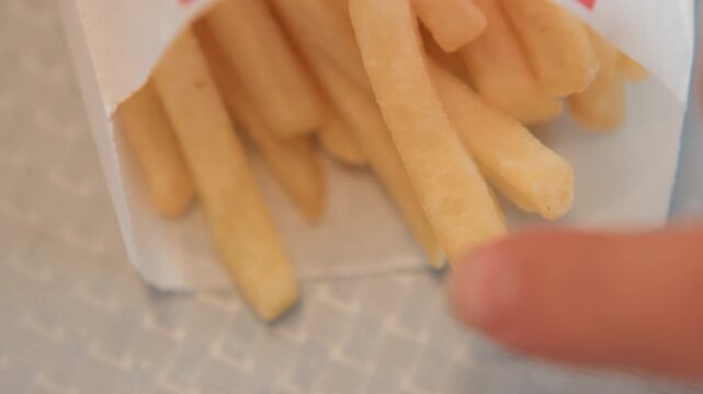 Close-up view of golden French fries in white paper container on patterned tabletop showing multiple small actions: fingers reaching and grasping fry, single fry slipping or sliding toward foreground