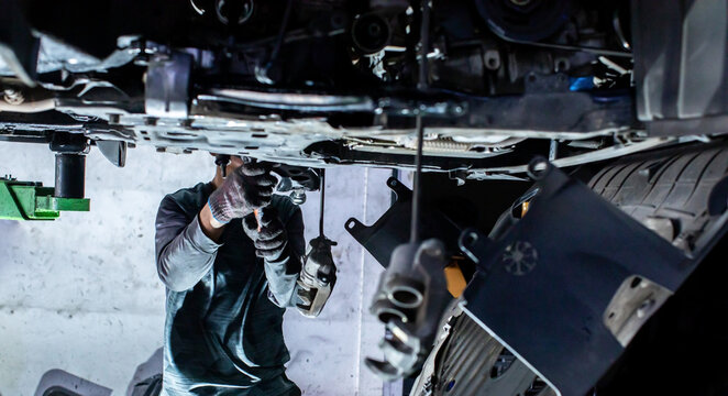 Selective focus to mechanic inspecting and repairing car underbody suspension components on a hydraulic lift inside an auto repair service workshop.