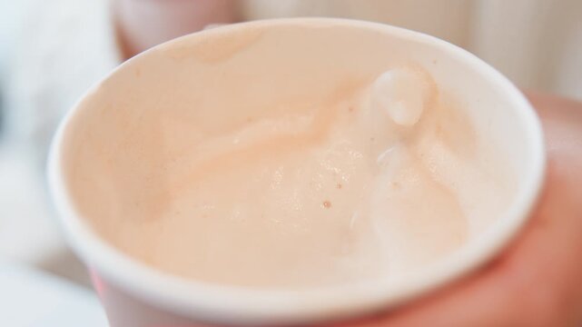 Top-front close-up of foam-covered hot latte in paper cup, creamy milk surface showing swirl marks, small bubbles, shallow dimples, and faint steam after recent fingertip touch, with warm soft light